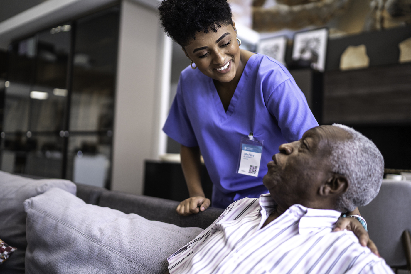 Female nurse in blue scrubs taking care of a senior man at home sitting on couch