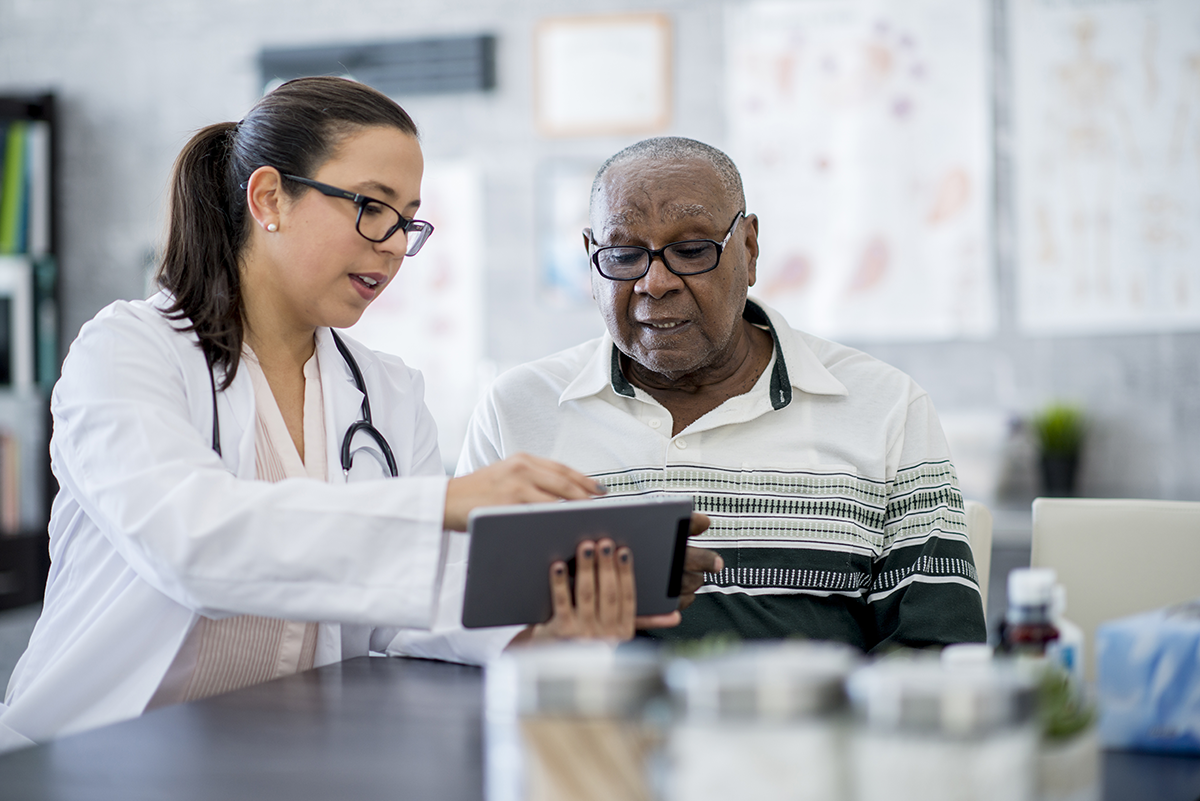 Female doctor with a tablet displaying information to an older man sitting at a table in doctor's office