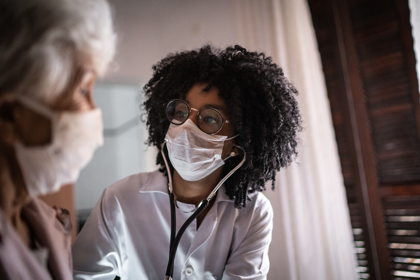 Doctor using stethoscope on a senior patient at home while both wear covid masks