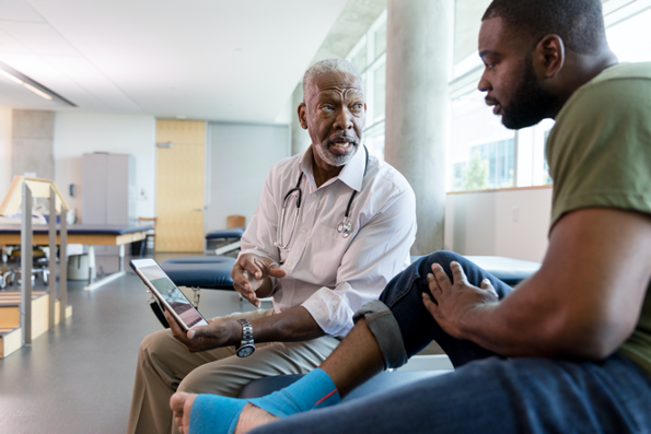 African-American doctor reading information from tablet to African-American patient sitting on bench in doctor's office