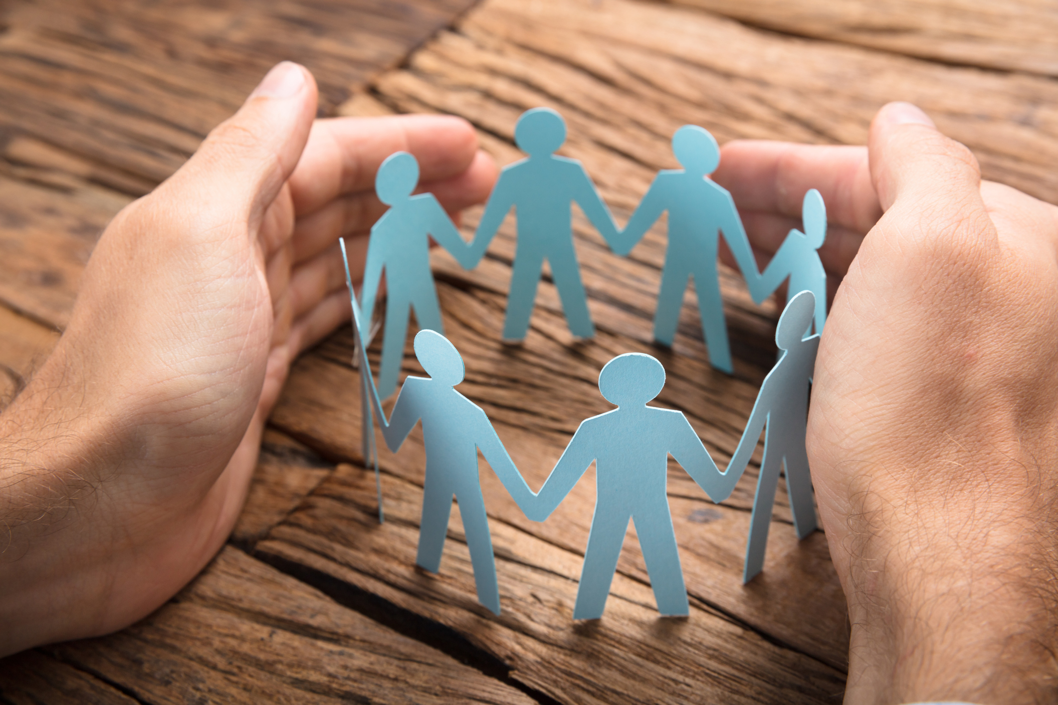 Image of two hands around a group of paper cut out people that are holding hands in a circle on a wooden table